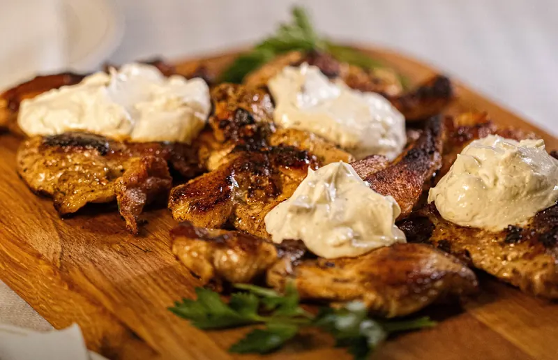 Grilled chicken pieces topped with spoonfuls of kajmak on a wooden board