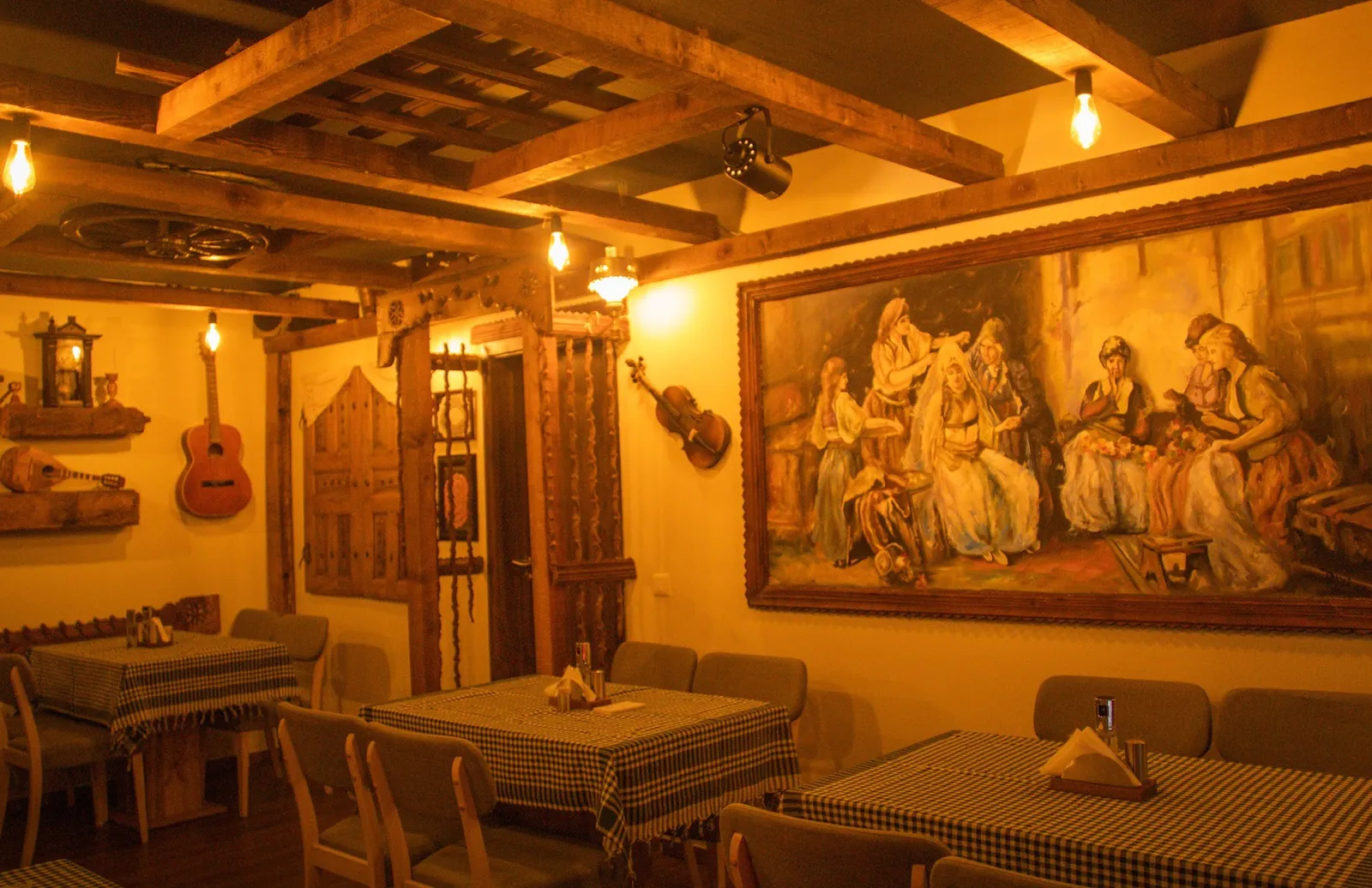 Warm-lit dining room with wooden beams, checked tablecloths and a large painting of a traditional gathering